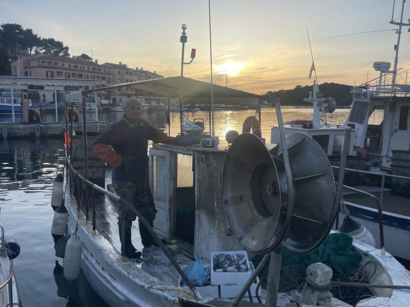 Fisherman in orange gloves on a small white boat at sunset, a crate of silver fish and a metal reel on deck