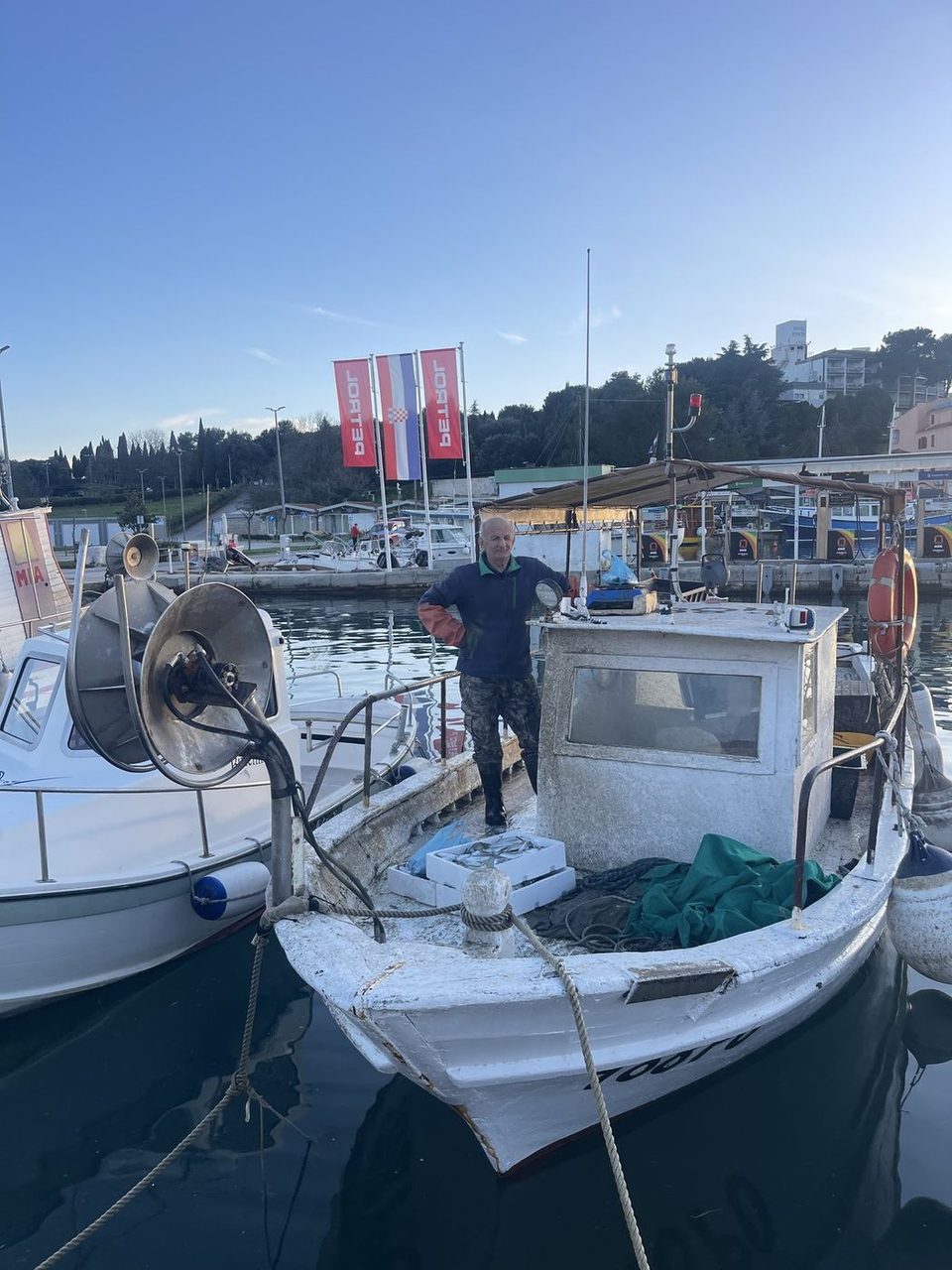 Weathered white fishing boat at a harbour quay in daylight, nets and crates on deck, coastal town behind